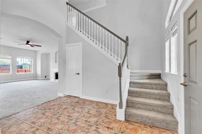 This photo showcases a bright entryway with tiled flooring and carpeted stairs leading to the second floor. The space opens into a large living area with carpet, a ceiling fan, and ample natural light from several windows. | Image 23