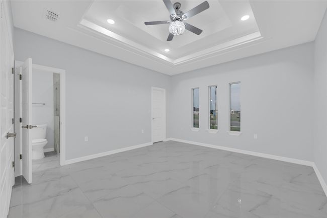 This is a bright, spacious bedroom with a modern design, featuring a tray ceiling with recessed lighting, a ceiling fan, and large glossy floor tiles. It includes three tall windows for natural light and an en-suite bathroom with a white door. | Image 28