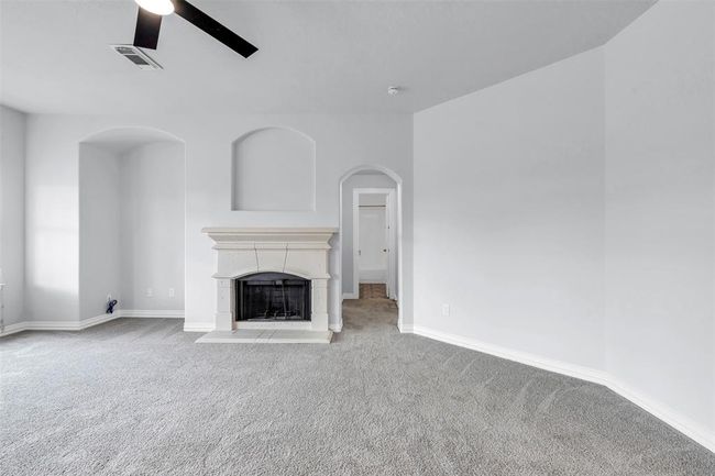 The room features a cozy fireplace set against a light-colored wall, complemented by a carpeted floor and an elegant ceiling fan. The space is bright and airy, with arched architectural details adding charm. A doorway leads to an adjacent area, enhancing the flow of the home. | Image 19