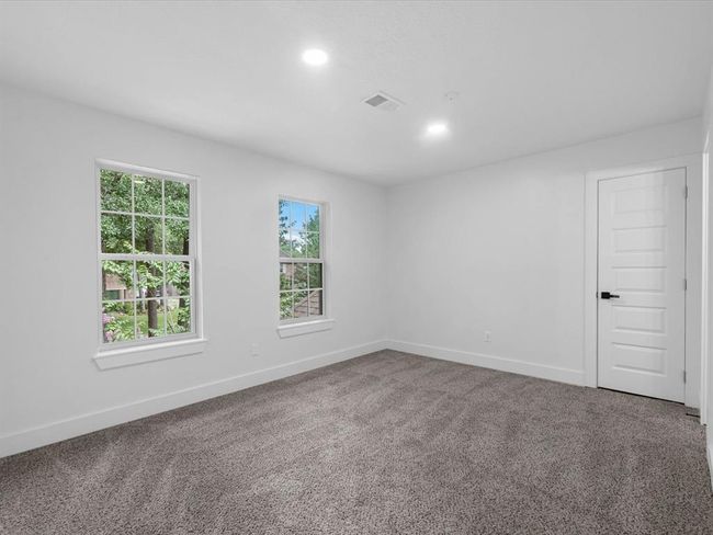 This photo shows a bright, spacious bedroom with neutral carpeting and white walls. It features two large windows offering natural light and outdoor views, along with a modern door. Ideal for creating a cozy and inviting personal space. | Image 26