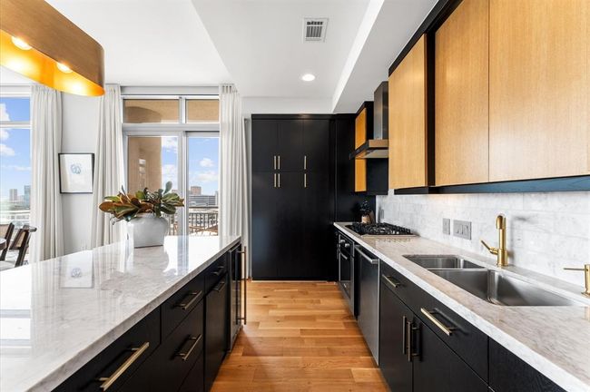 This kitchen features modern design with sleek black and wood cabinetry, marble countertops, and gold hardware. It includes a Bosch gas stove, double sink, Frigidaire refrigerator and large windows that provide ample natural light. The space opens to a view of the cityscape, enhancing its contemporary appeal. | Image 4