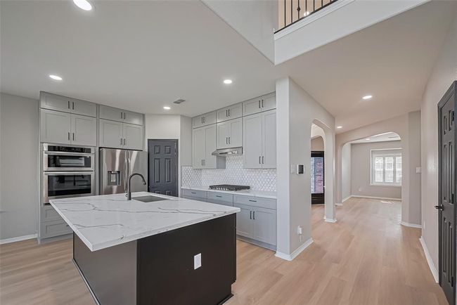 This modern kitchen features sleek gray cabinetry, stainless steel appliances, and a large marble island with a built-in sink. Open layout with bright lighting and an adjacent living space. | Image 11