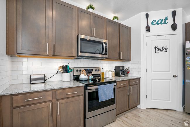 Kitchen with light wood finished floors, dark brown cabinetry, decorative backsplash, light stone countertops, and appliances with stainless steel finishes | Image 7