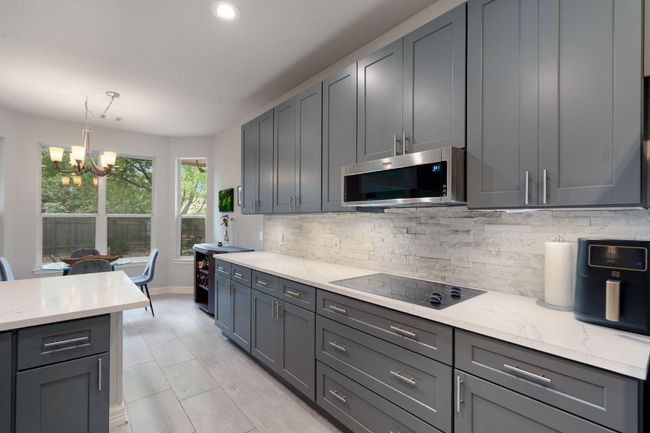 Kitchen with black electric cooktop, stainless steel microwave, tasteful backsplash, a chandelier, and light stone countertops | Image 14