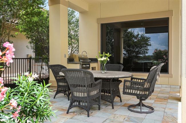 This photo features a cozy covered outdoor patio with a stone tile floor, and a built-in grill. The area is enhanced by lush greenery and bright flowers, offering a pleasant space for outdoor dining and relaxation. | Image 46