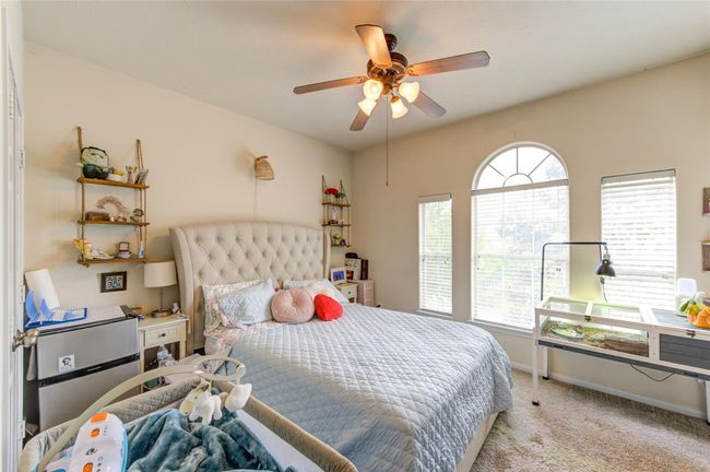 Cozy bedroom with a plush tufted bed, large arched windows for natural light, ceiling fan, and carpeted flooring. Shelving adds storage and charm, while the room remains bright and inviting. | Image 30