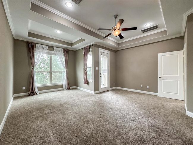 This spacious room features a tray ceiling with recessed lighting, a ceiling fan, neutral carpet, and large windows with elegant drapes, providing ample natural light. A door leads to an outdoor area, and the overall design is modern and inviting. | Image 25