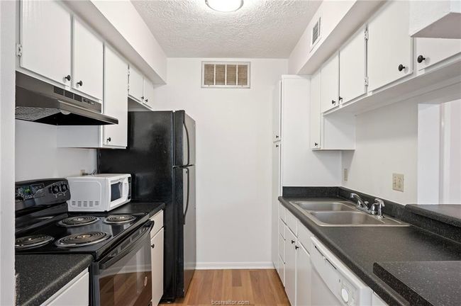 Kitchen featuring white appliances, light wood-type flooring, under cabinet range hood, white cabinetry, and dark countertops | Image 5