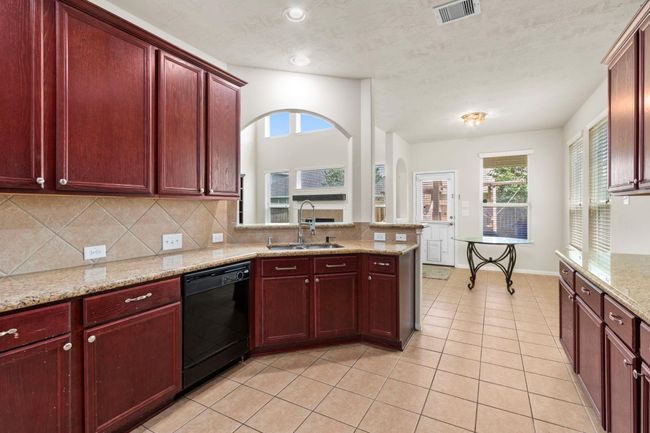 This bright kitchen features rich wood cabinetry, granite countertops, and a central island with a sink. It opens to a dining area with large windows and tile flooring, creating a spacious and inviting atmosphere. | Image 9