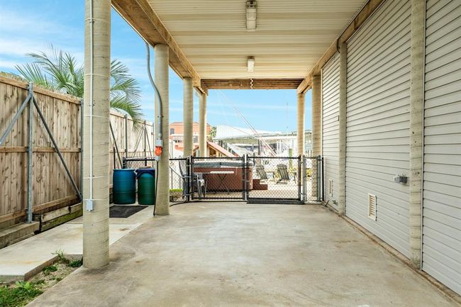 This photo shows a covered outdoor area with a concrete floor, surrounded by a wooden fence and metal gate. The space is supported by sturdy columns and features a view of a backyard with seating. It’s ideal for parking or outdoor activities. | Image 46