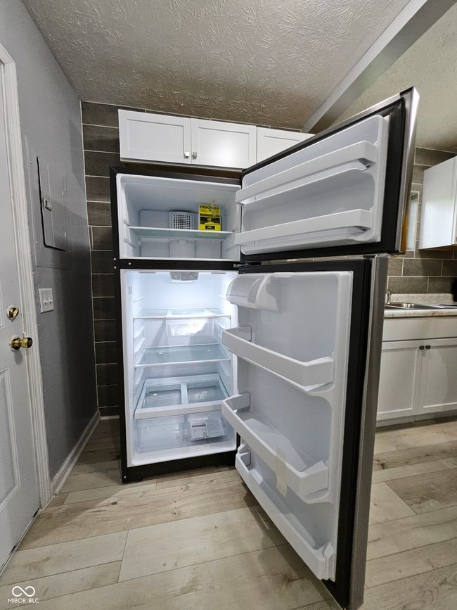 kitchen view of light wood-type flooring, a textured ceiling, refrigerator, and white cabinets | Image 12