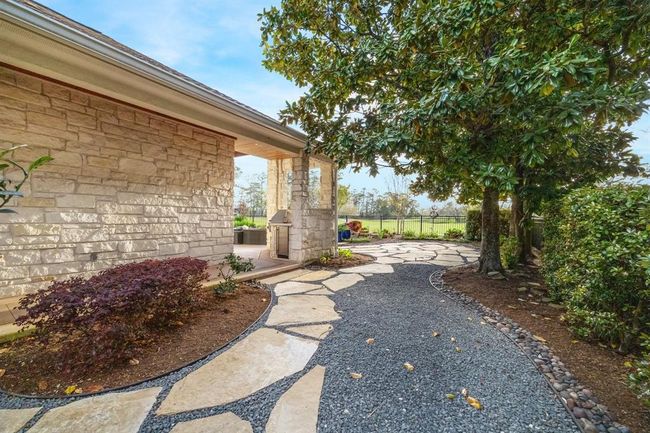 This photo showcases a charming backyard with a stone pathway leading to a patio area. The space is bordered by lush greenery and mature trees, offering a peaceful outdoor retreat. The stone exterior of the house complements the natural setting. | Image 37