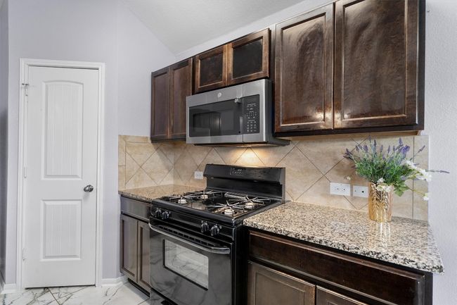Kitchen featuring black range with gas cooktop, stainless steel microwave, dark brown cabinets, light stone countertops, and lofted ceiling | Image 10