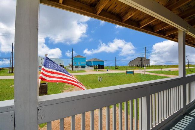 This photo showcases a view from a covered deck, featuring a neatly maintained lawn and distant houses, including a striking blue one. An American flag adds a patriotic touch, and the scene is set against a backdrop of a bright, partly cloudy sky. | Image 5