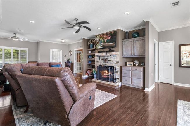 This inviting living room features cozy brown leather seating, a stone fireplace, and built-in shelving. The space is complemented by hardwood floors, ceiling fans, and ample natural light from large windows, creating a warm and welcoming atmosphere. | Image 8