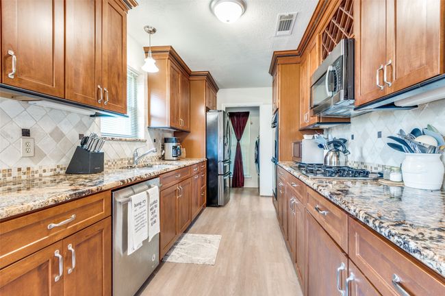 Kitchen with stainless steel appliances, brown cabinetry, light wood-style flooring, decorative backsplash, and light stone counters | Image 6