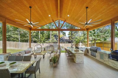 This is a spacious covered patio with a high wooden ceiling, multiple ceiling fans, and a view of the backyard. It features a built-in grill, a long dining table, and comfortable lounge areas. | Image 1