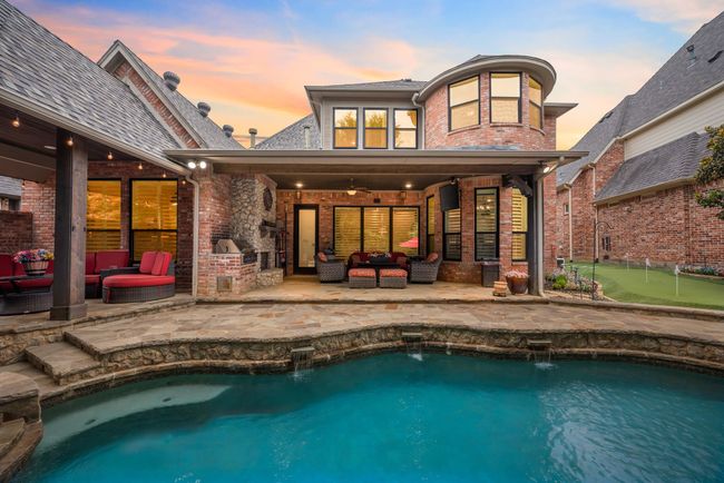 Rear view of property featuring 2 outdoor living areas that include a fireplace and built in grill. The pool has multiple water features and to the side is a 2 hole putting green with synthetic turf. Outdoor lighting enhances the evening views. | Image 39
