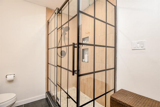 Modern bathroom with a sleek glass shower featuring black grid framing and wood-textured tile. The space is bright with a minimalist design, including a wall-mounted toilet and light switch panel. | Image 30