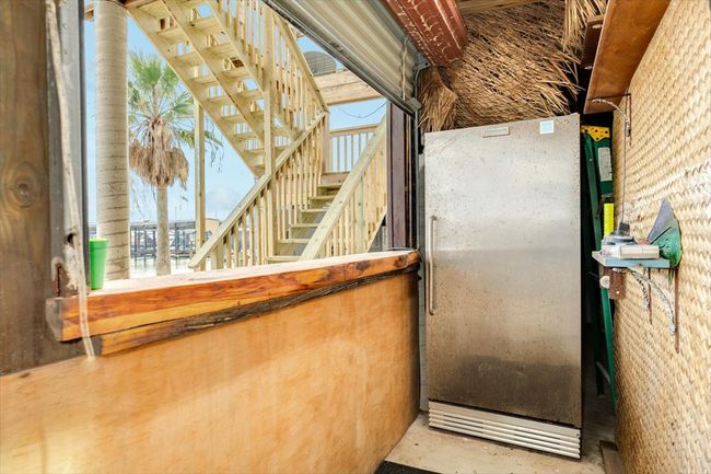 This photo features an outdoor area with a rustic vibe, including a metal refrigerator and wooden bar. A staircase leads to an upper level, and there's a view of a palm tree and waterfront in the background. | Image 43