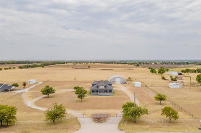 This aerial image shows the spacious property with a single-family home, featuring a large front yard, an expansive workshop and other outbuildings, set amid expansive open fields, offering unobstructed views, privacy, and rural charm. Property includes TWO 500 gallon septic tanks. Rhome is an easy commute from Dallas area and only 12 miles from Texas Motor speedway so great primary residence, weekend getaway, or EXCELLENT investment opportunity! | Image 1