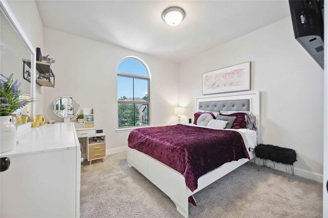 This cozy bedroom features a plush carpet, a large arched window for natural light, and a stylish white and purple color scheme. The room includes a comfortable bed, a dresser, and modern decor, creating a welcoming and relaxing atmosphere. | Image 31