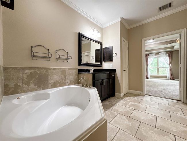 This is a spacious bathroom featuring a large soaking tub, a sleek vanity with a framed mirror, and neutral tile flooring. It opens to a bright bedroom with carpet and large windows, creating a seamless flow between the spaces. | Image 37