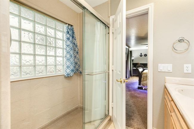 This photo features a bathroom with a glass shower, textured glass block window, and a view into a carpeted bedroom. The bathroom has light-colored walls and a countertop, creating a bright and inviting space. | Image 16