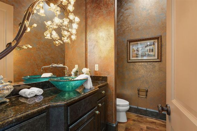 This elegant powder room offers added convenience for guests, featuring a Venetian plastered metallic wall finish, a unique glass vessel sink, and a luxurious floral light fixture. It includes dark granite countertops and wood flooring, creating a sophisticated atmosphere. | Image 18