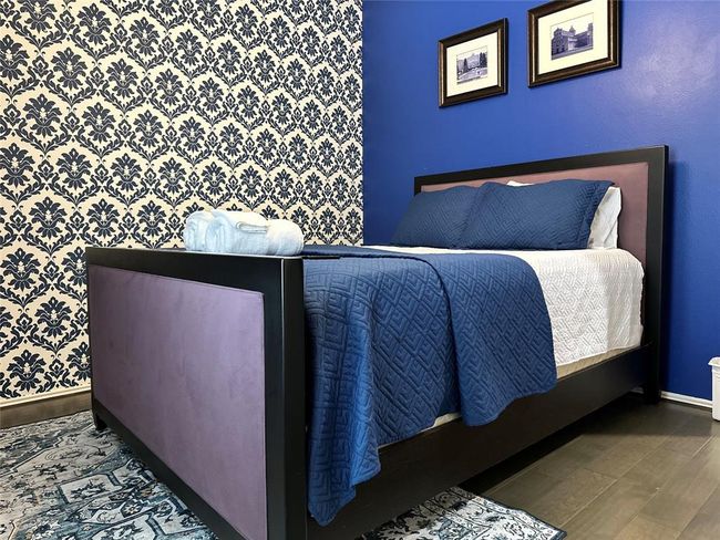 This bedroom features a stylish design with a blue accent wall and a patterned wallpaper. It includes a modern bed with deep blue bedding, complemented by framed artwork. The wood flooring and a decorative rug add warmth and character. | Image 12