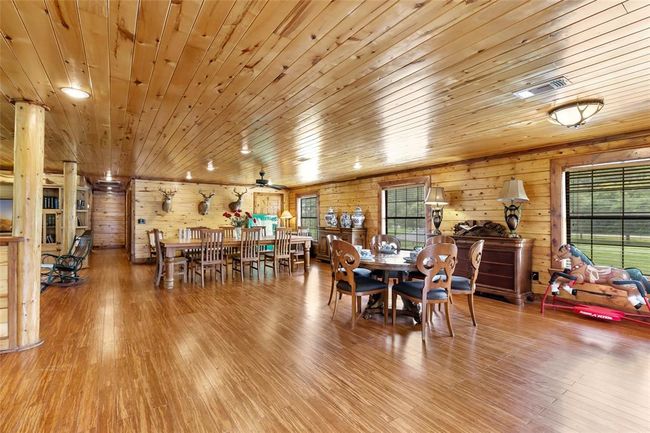 This spacious, rustic dining and living area features warm wood paneling, hardwood floors, and ample natural light. The room is equipped with large dining tables, decorative lighting, and classic wooden furniture, creating a cozy, inviting atmosphere. | Image 48