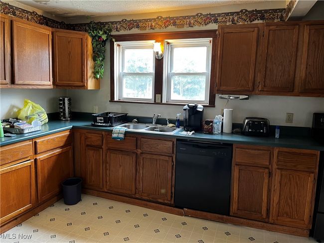 Kitchen featuring black dishwasher, light tile patterned flooring, dark countertops, brown cabinetry, and a sink | Image 9