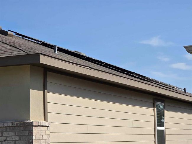 This photo shows the side view of a house with beige siding and a shingled roof. There are solar panel mounts on the roof, a brick corner, and a window on the right. The sky is clear and blue. | Image 25