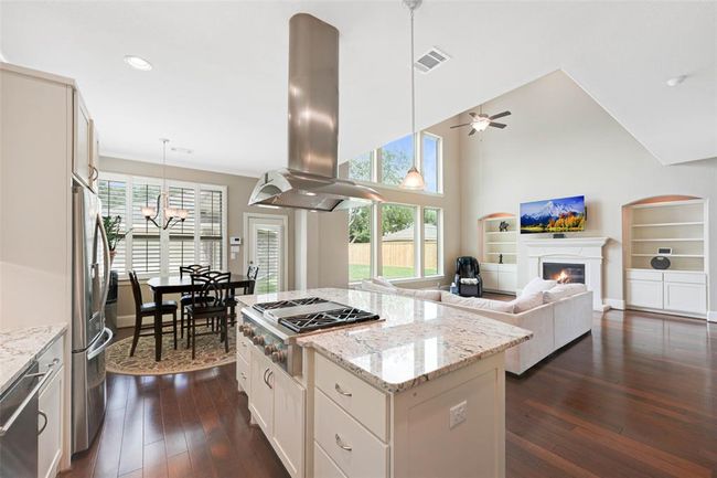 Open-concept layout with seamless flow from kitchen to breakfast area and living room. Features include a large granite island with gas cooktop, vaulted ceilings, built-in shelving, fireplace, and oversized windows overlooking the backyard—perfect for comfortable everyday living. | Image 12