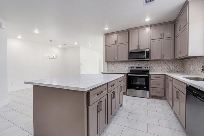 This photo showcases a modern kitchen with sleek cabinetry, stainless steel appliances, and a spacious island. The neutral color palette, combined with the stylish backsplash and tile flooring, creates a clean and inviting atmosphere. The open layout leads to a dining area with a contemporary chandelier. | Image 13
