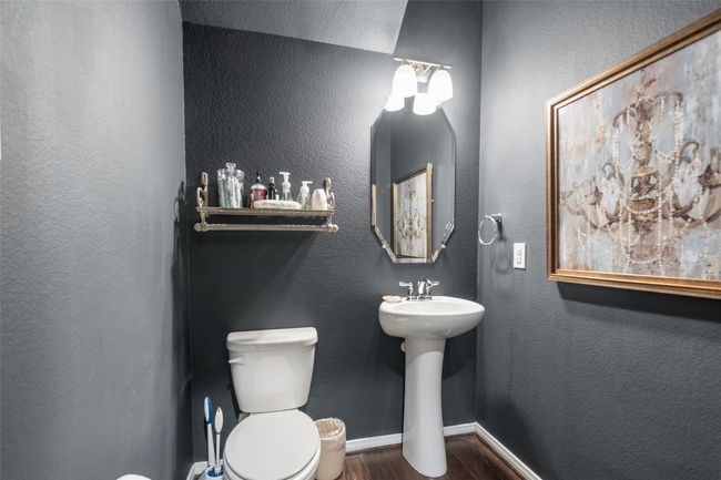 This is a compact, stylish half-bathroom with dark gray walls and wood flooring. It features a pedestal sink, a toilet, and a decorative mirror. A wall-mounted shelf above the toilet provides space for toiletries. | Image 25