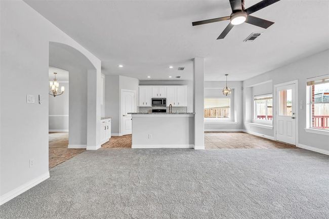 This photo showcases a bright, open-concept living area with a modern ceiling fan and neutral carpeting. The space seamlessly connects to a kitchen featuring white cabinetry, granite countertops, and stainless steel appliances. Large windows and a glass door provide ample natural light and access to the backyard. | Image 9