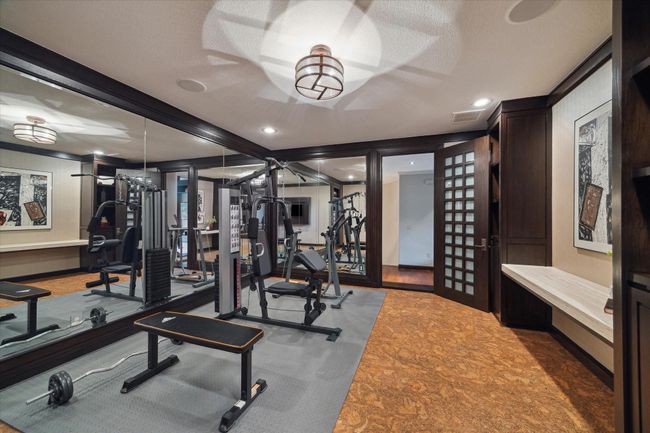 This well-appointed fitness room offers a motivating space to focus on health and wellness. Mirrored walls enhance the sense of openness and light, while sound dampening cork flooring ensures comfort and safety during training. Built-in shelving and a desk nook provide convenient storage and organization, and the adjacent flex room creates seamless access for post-workout relaxation. Through the custom built-in bookshelf, you’ll find a hidden room currently being used as an art studio. | Image 38