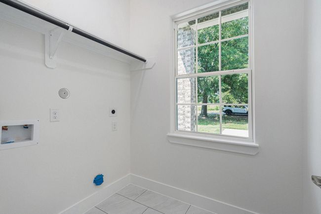 This photo shows a bright laundry room with a window providing natural light. It features hookups for a washer and dryer, tiled flooring, and a hanging rod for clothes. | Image 31