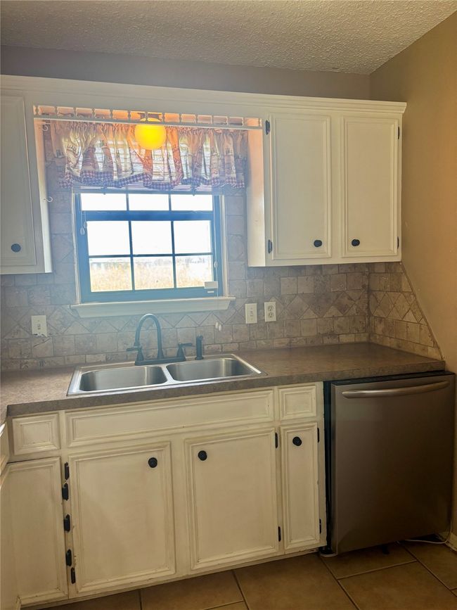 Kitchen with stainless steel dishwasher, light tile patterned floors, decorative backsplash, white cabinets, and a textured ceiling | Image 10