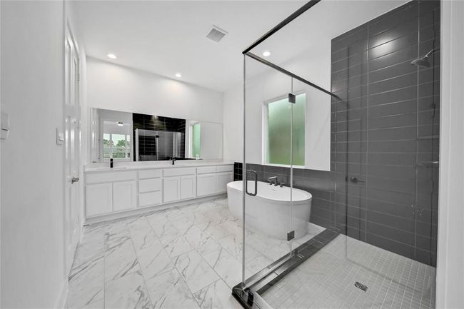 This contemporary bathroom features a sleek freestanding tub, elegant black tile accents, and a stylish vanity. The space is complemented by artistic wall decor and a frosted window for privacy and natural light. | Image 27