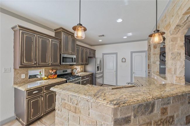 Kitchen featuring appliances with stainless steel finishes, dark brown cabinets, crown molding, backsplash, and light stone countertops | Image 13