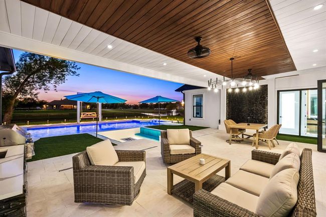 This stunning outdoor oasis boasts a luxurious covered patio with rich wood-paneled ceiling accents, recessed lighting, and ceiling fans for year-round comfort. A sleek built-in grill and prep area enhance the convenience of the outdoor kitchen. Overlooking a contemporary pool with vibrant lighting, spa, and in-pool lounge, the space is framed by open views and elegant landscaping—perfect for relaxing or entertaining in style. | Image 15
