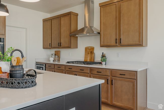 Kitchen with stainless steel gas cooktop, brown cabinets, wall chimney range hood, and light countertops | Image 7