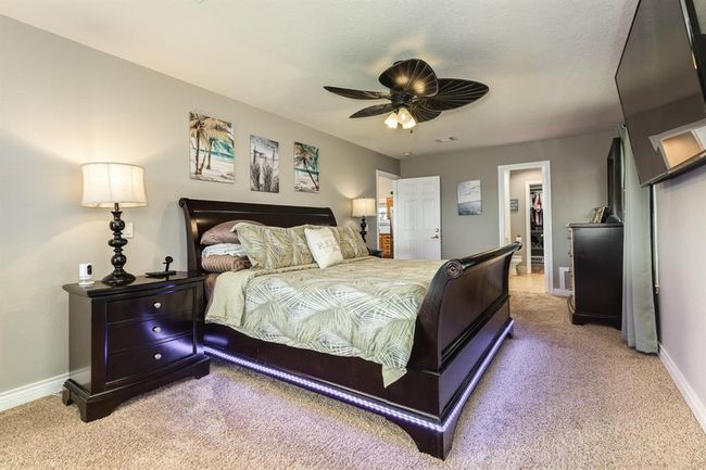 This spacious bedroom features a cozy carpeted floor, a stylish dark wood bed with under-bed lighting, and a ceiling fan. An en-suite bathroom and walk-in closet offer convenience, while wall art adds a touch of elegance. | Image 14