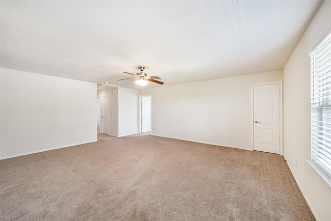 This spacious upstairs game room features neutral carpeting, a ceiling fan, and ample natural light from a large window. It has clean white walls and direct access to a hallway and adjacent rooms through white doors. | Image 18