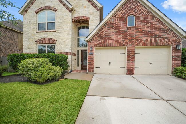 This charming two-story home features a brick and stone facade, a three car garage, and a well-maintained front lawn with shrubs. The arched windows and entryway add a touch of elegance to the exterior. | Image 4
