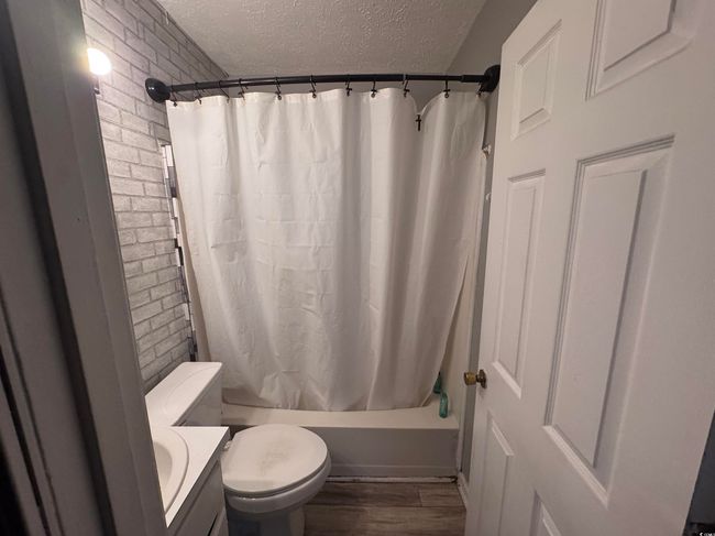 Full bath featuring a textured ceiling, brick wall, toilet, shower / tub combo with curtain, and wood finished floors | Image 4