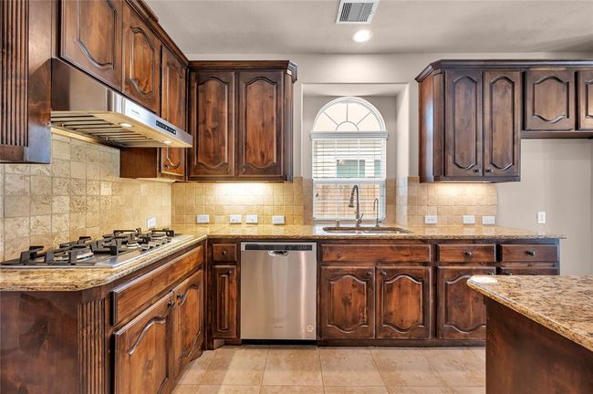 This kitchen features rich wooden cabinetry, granite countertops, a gas stove, and a stainless steel dishwasher. It has a tile backsplash and flooring, with a charming arched window above the sink for natural light. | Image 13