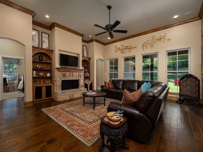 Living area featuring dark wood-style floors, a stone fireplace, a ceiling fan, ornamental molding, and built in features | Image 6